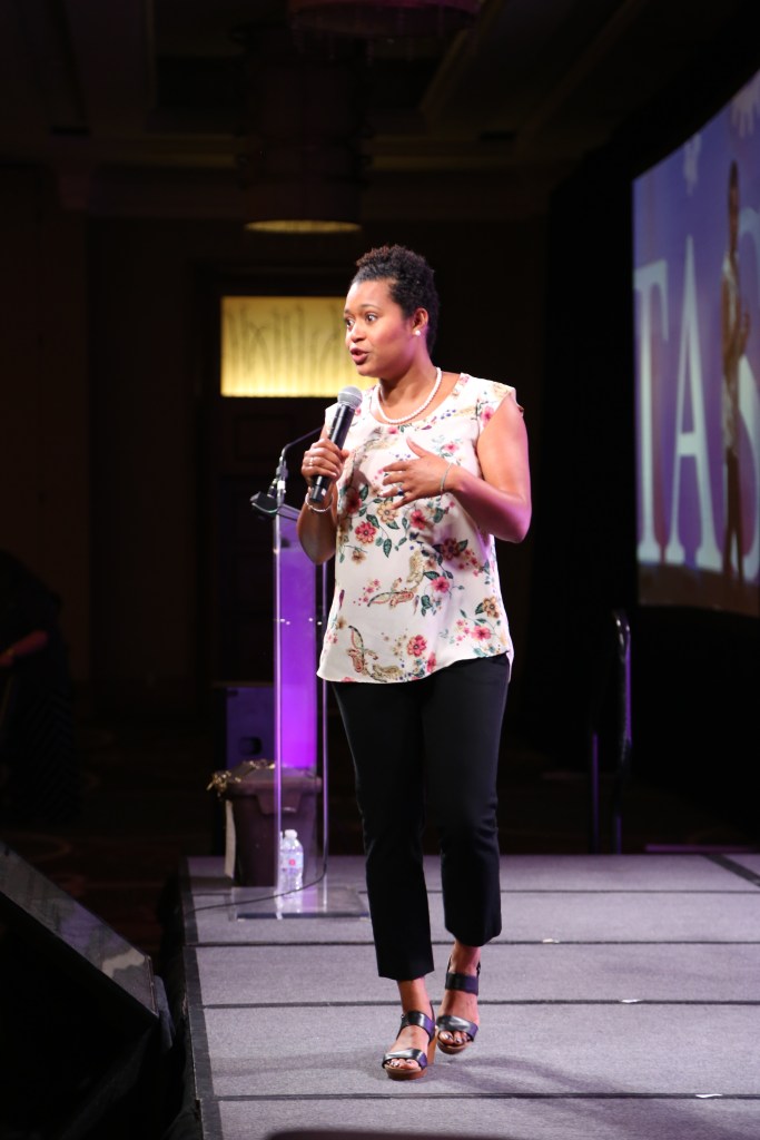 Kori Hamilton Biagas walking across stage talking into a microphone giving a speech. 